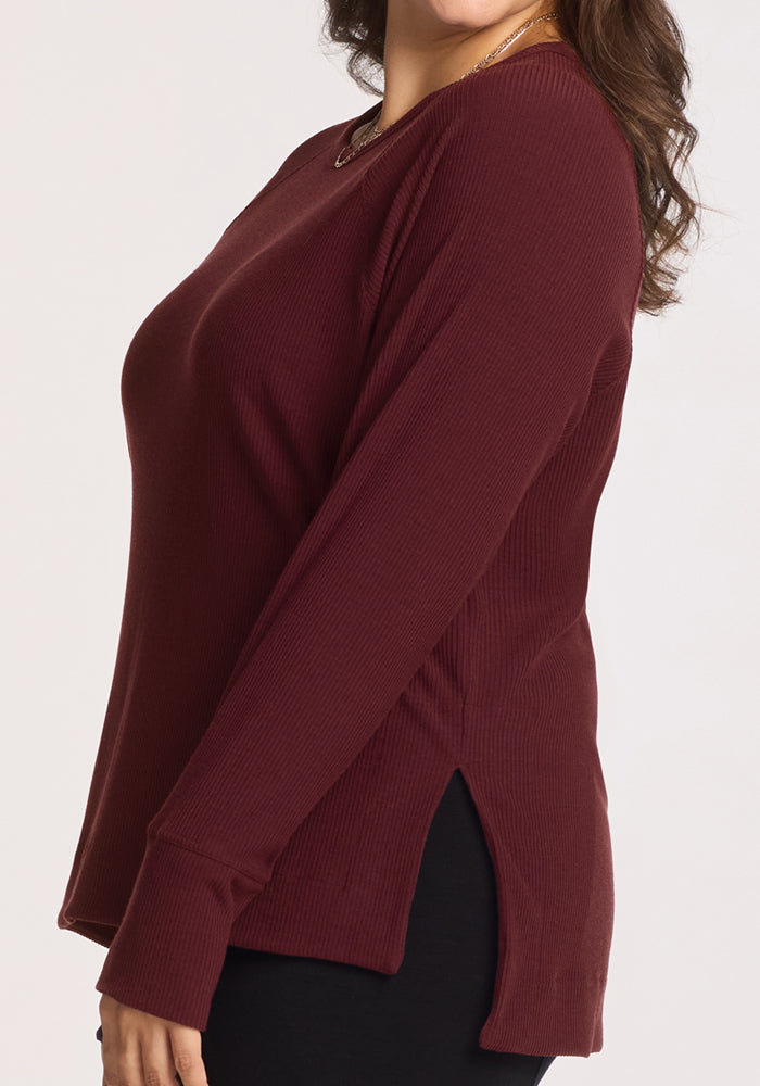 A person is shown from the side against a plain light background wearing the Woolx Ainsley Sweater in Toasted Garnet, featuring long sleeves, side slits, and a ribbed merino wool design.