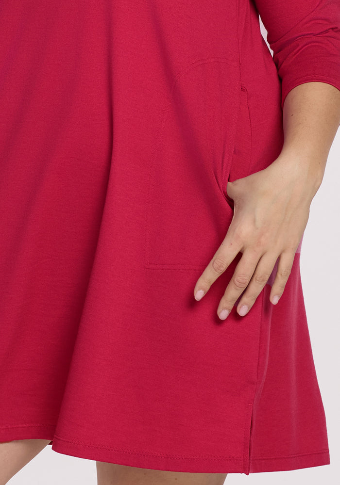 Someone wears the Woolx Rissa A Line Dress in Poppy Spark—a long-sleeved, bright pink Merino wool dress with a side pocket, perfect for cozy fall-winter layering. Only part of their arm and hand are visible in the image.