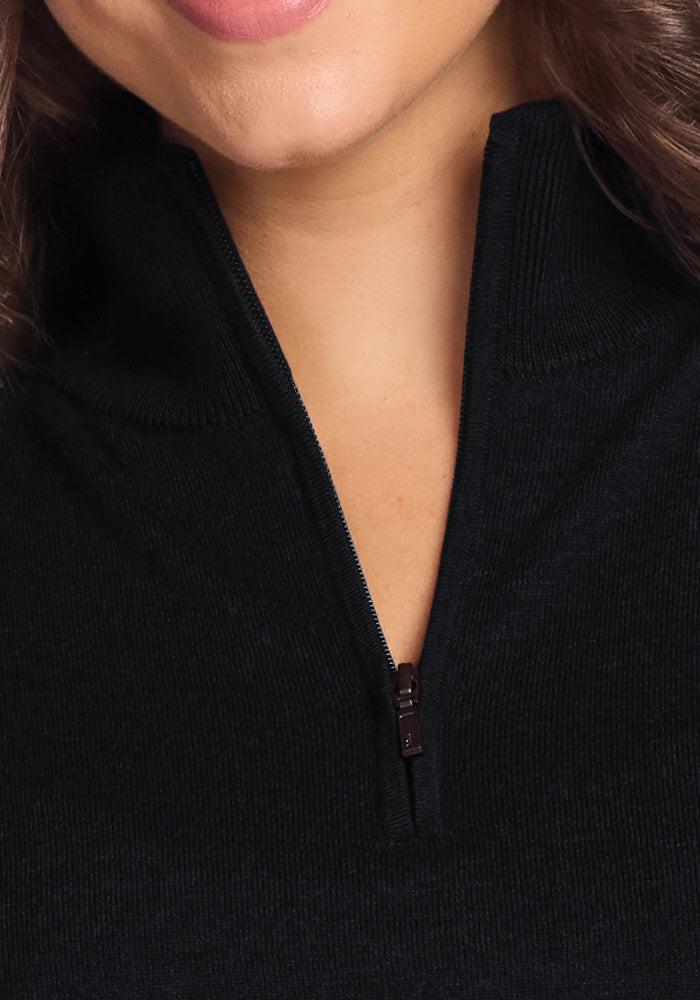 Close-up of a woman smiling, with curly brown hair, wearing the Woolx Collins Half-Zip Sweater in black. The merino wool sweater’s zipper is partially open, revealing her lower face and neck.