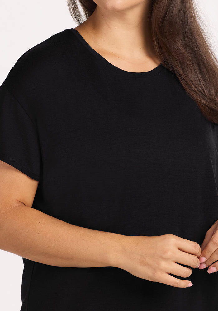 A woman with long brown hair wears the Woolx Billie Lounge Tee in black, shown from shoulders to mid-torso with her hands gently clasped in front.