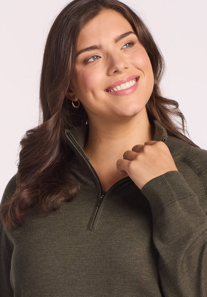 A woman with long brown hair and gold hoop earrings smiles upward, wearing the Woolx Collins Half Zip Sweater in Pine Needle. She has one hand raised near the sweater's zip-up collar.