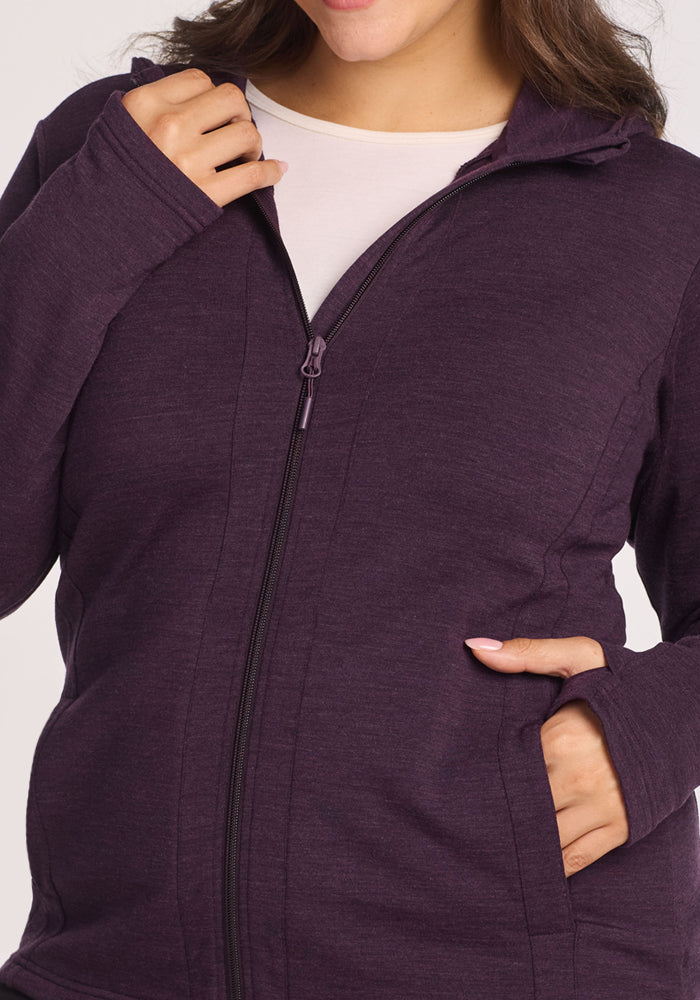 A woman wears a Woolx Zoey Zip Up Hoodie in Deep Plum over a white top, one hand on the zipper and the other in the pocket. Her face is partially out of frame.