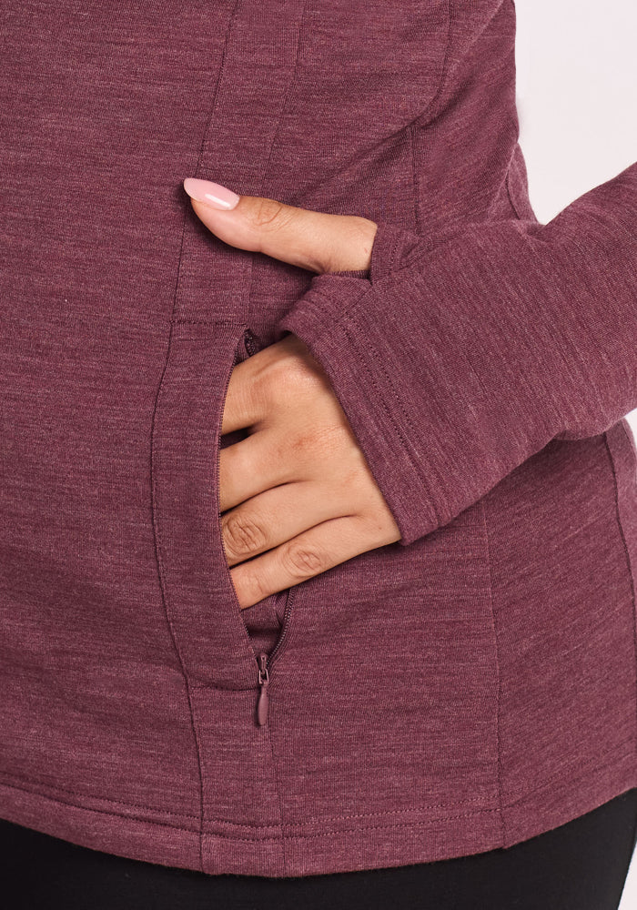 A close-up of a hand, with light pink nail polish, partially in the zippered pocket of the Woolx Finley Zip Up Sweatshirt in Grape Harvest Melange. The long sleeve features a thumbhole and is made from odor-resistant fabric.