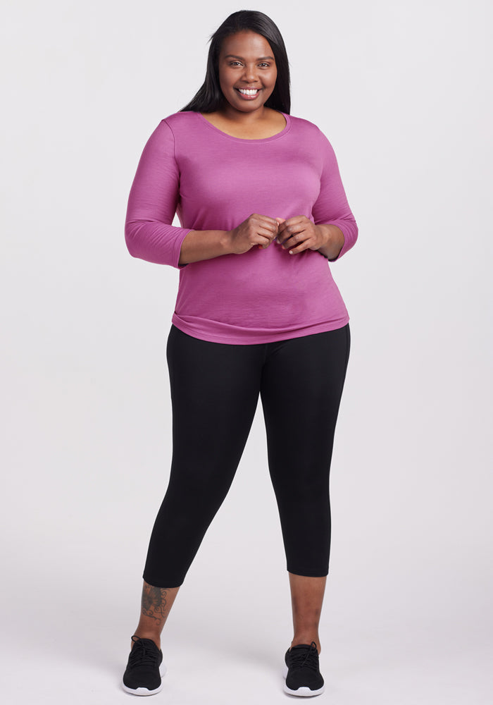 A woman with long dark hair smiles while wearing the Woolx Jenny 3/4 Sleeve Crew Neck in Purple Wine, paired with black leggings and sneakers. She stands against a light, plain background.