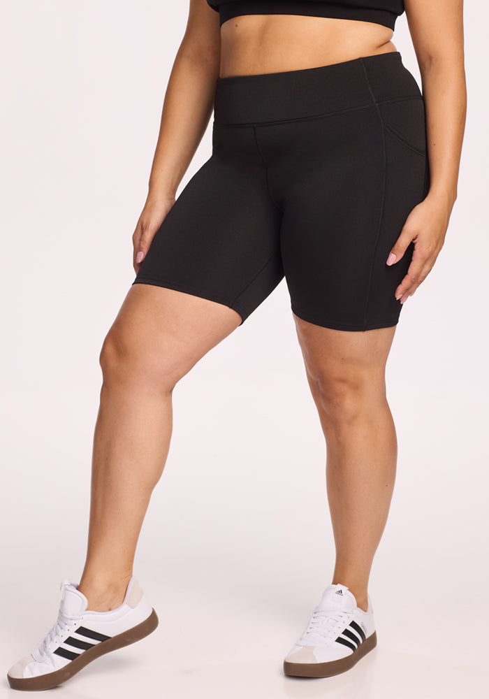 A person wears Woolx Merino Flex® Dani Bike Shorts in black, paired with a matching sports bra and white sneakers with black stripes. Only their lower torso and legs are shown against a plain white background.