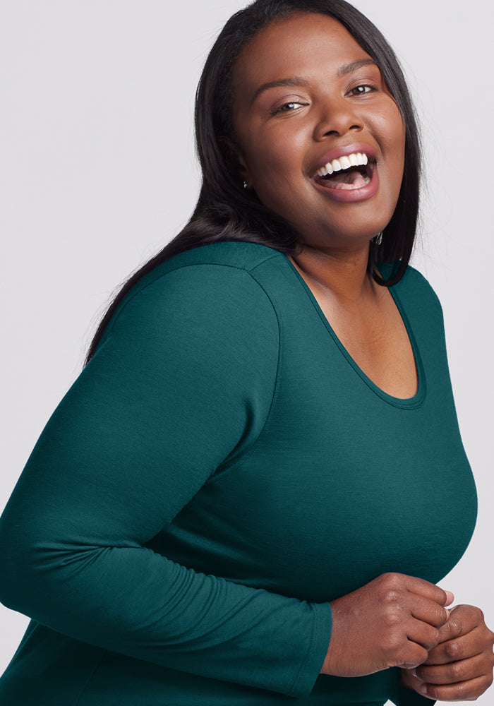 A woman with long dark hair smiles, wearing the Woolx Remi Long Sleeve T-Shirt in Riverbank Green against a plain light background.