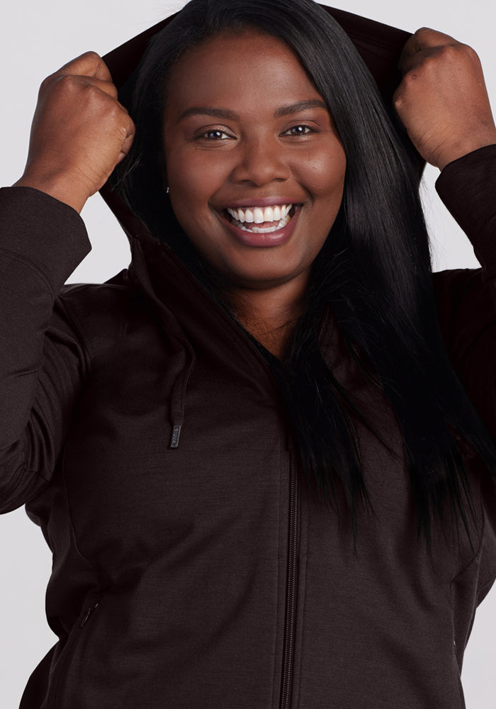 A smiling woman with long dark hair pulls up the hood of the Woolx Ryann Hoodie in French Roast, looking directly at the camera against a plain light background.