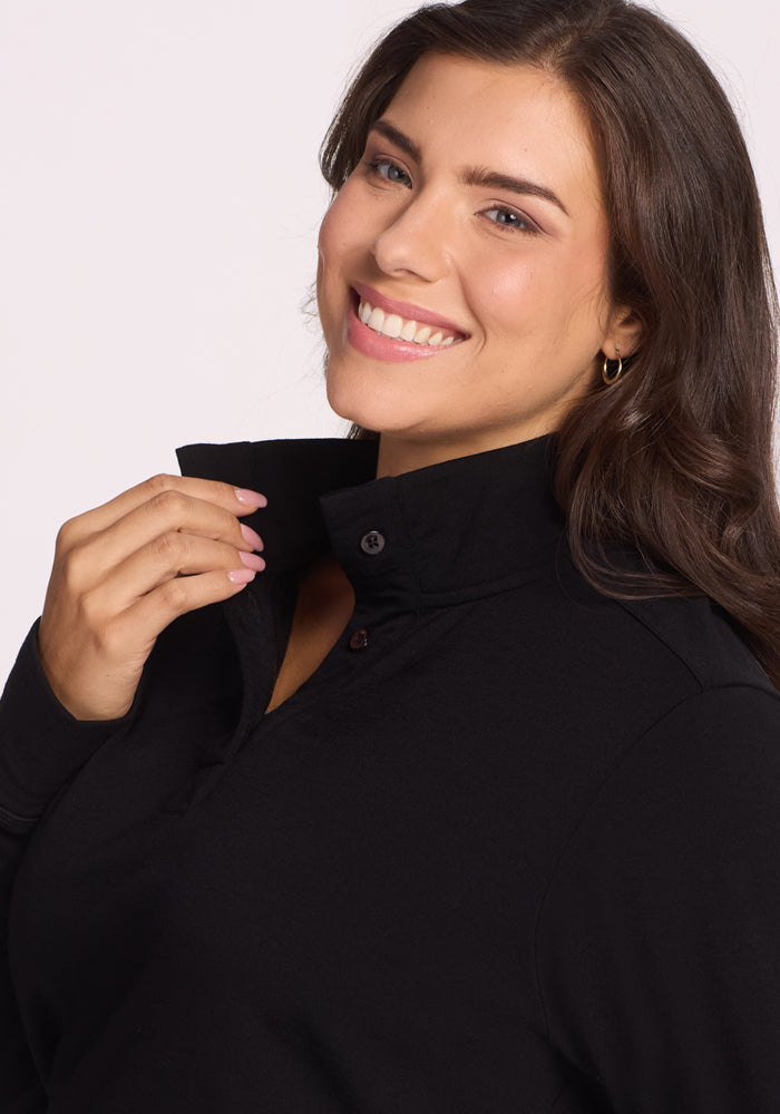 A woman with long brown hair smiles while holding the collar of her Woolx Annie 3 Button Pullover in black. She wears light makeup, gold hoop earrings, and light pink nail polish against a plain, light-colored background.