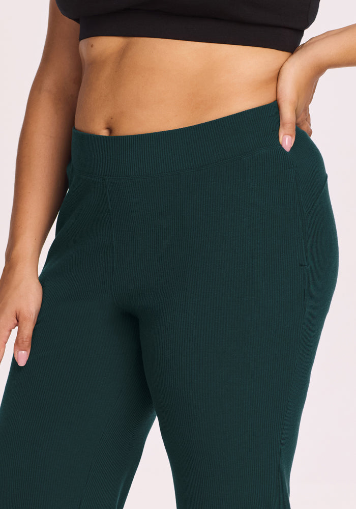 Someone models Woolx’s Luca Ribbed Pants in Deep Timber, styled with a black cropped top. Only the waist and upper legs are shown, with one hand resting on the hip.