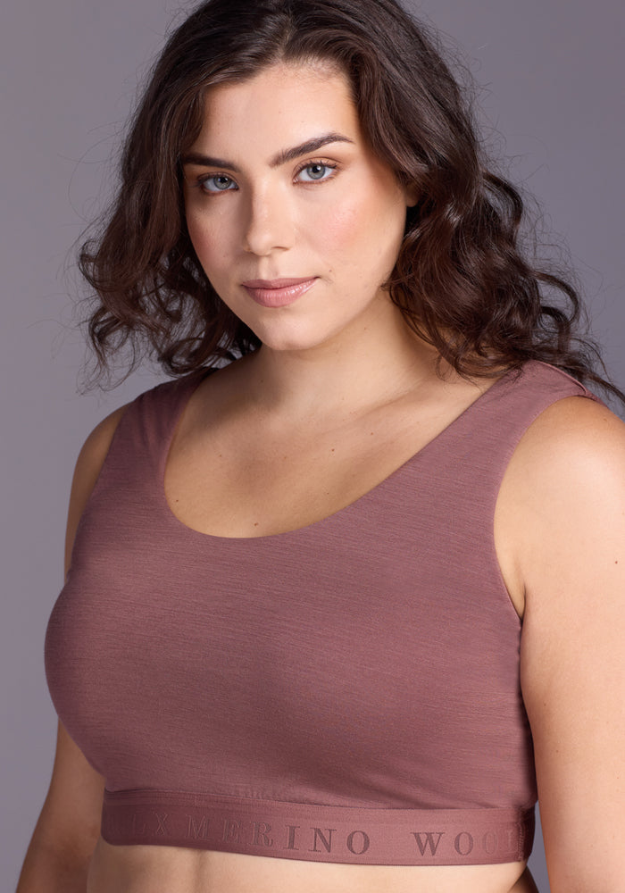 A woman with wavy brown hair wears the Woolx Aubrey Lounge Bra in Nutmeg, featuring a "MERINO WOOL" logo band, as she stands confidently against a gray background.