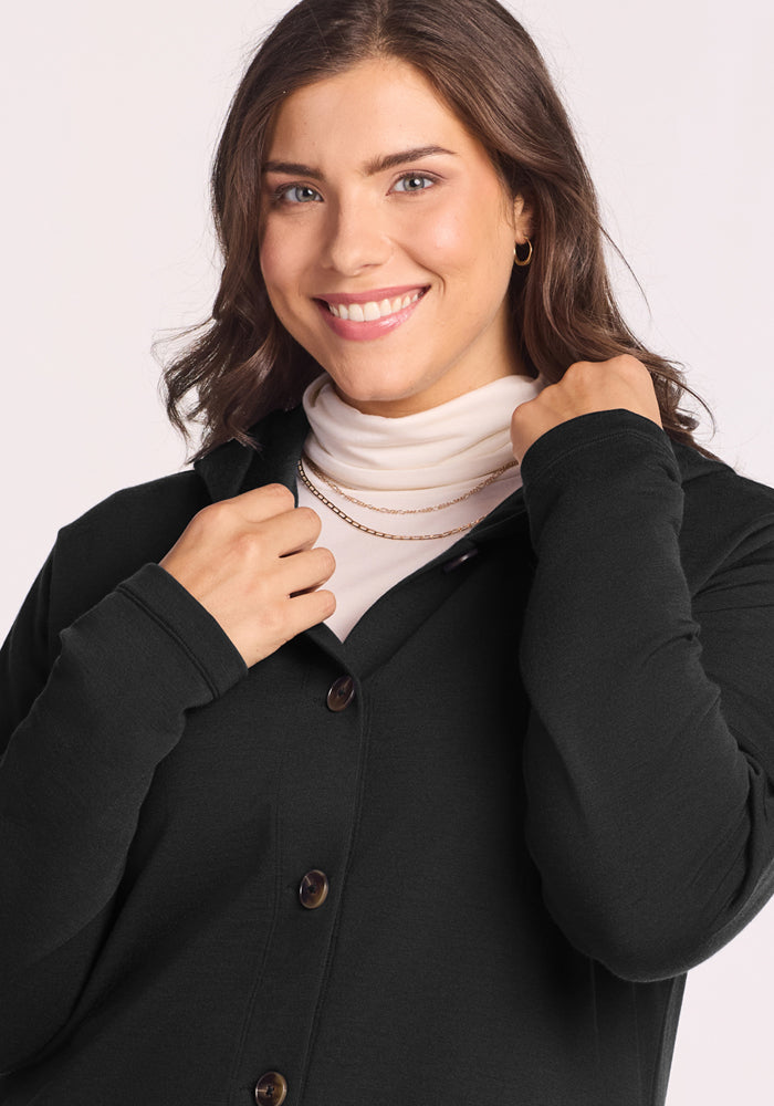 A woman with wavy brown hair smiles, holding the collar of her Woolx Edna Hooded Cardigan in black. She layers it over a white turtleneck and accessorizes with gold hoop earrings and a layered gold necklace against a light background.