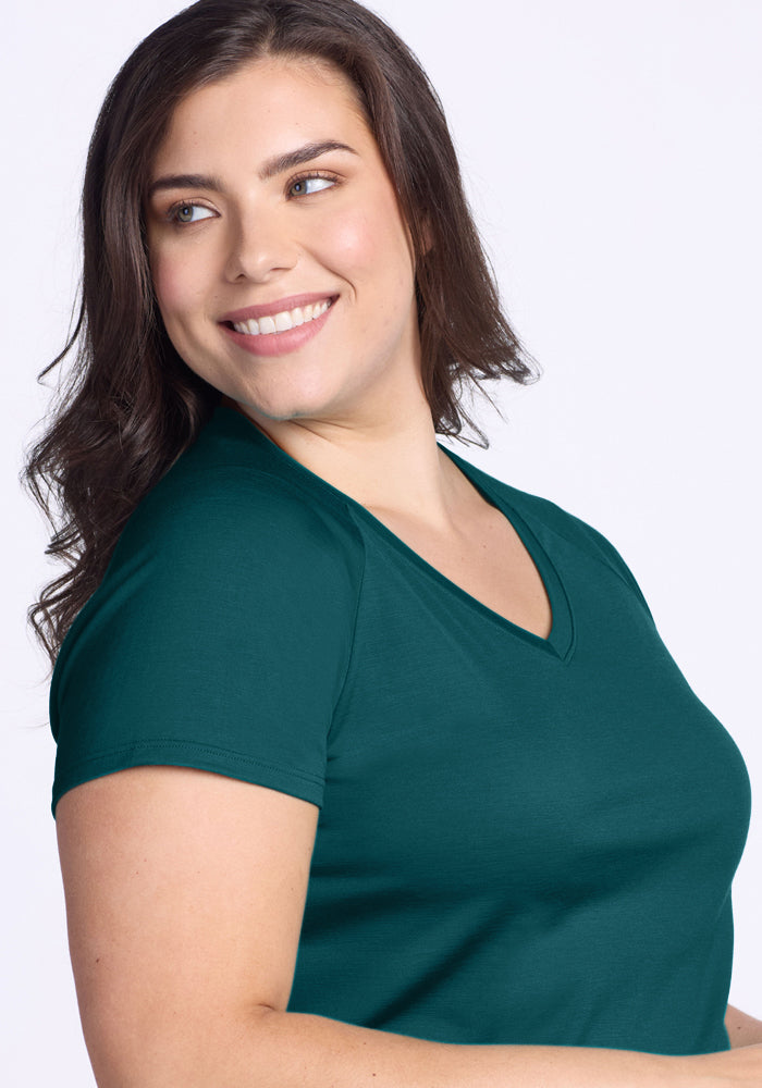 A woman with long brown hair smiles and looks to the side, wearing the Woolx Mia V Neck in Riverbank Green—a lightweight, odor-resistant Merino wool short-sleeve tee—set against a light background.