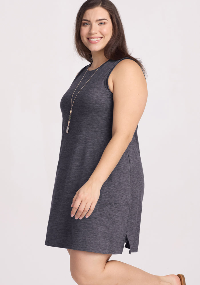 A woman with long brown hair smiles while standing sideways against a light background, wearing the Woolx Clara Dress in Cinder Melange and a long pendant necklace.