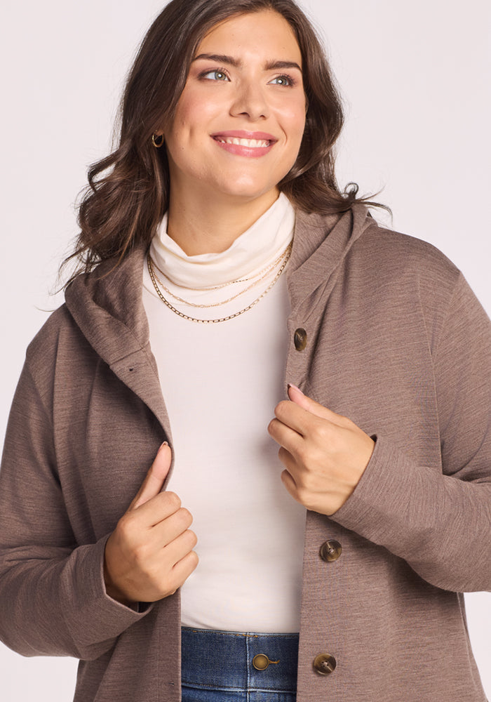 A woman with wavy brown hair smiles while modeling the Woolx Edna Hooded Cardigan in Simply Taupe over a white turtleneck and blue jeans, accessorized with gold necklaces, holding the lapel against a light background.