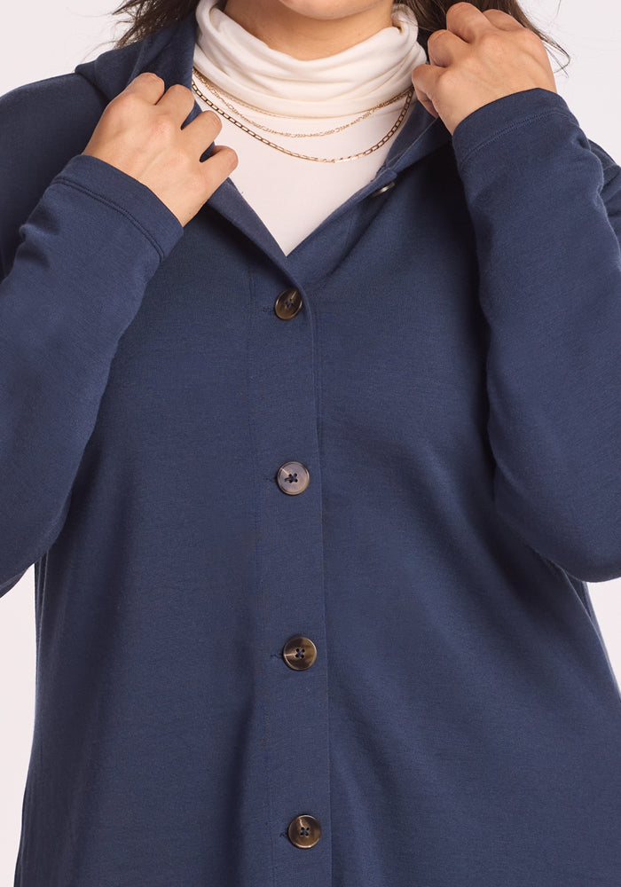A person wears the Woolx Edna Hooded Cardigan in Deep Navy over a white turtleneck, accessorized with two gold chain necklaces, adjusting the collar—ideal for cold-weather layering.