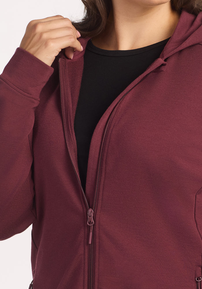 A person wears the Woolx Darby Zip Up Hoodie in Toasted Garnet over a black top, shown from shoulders to waist, with one hand holding the zipper near the neck. The hoodie is partially unzipped for a casual look.