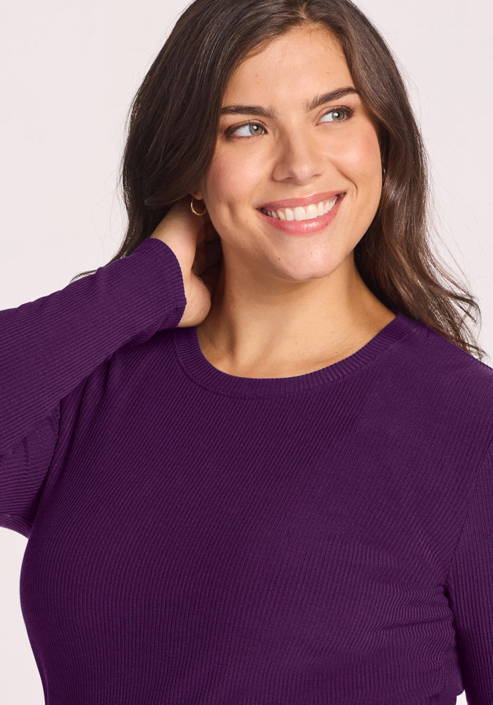 A woman with long brown hair smiles and looks to the side, her hand behind her neck, wearing the Woolx Hadley Ribbed Crew in Majestic Purple.