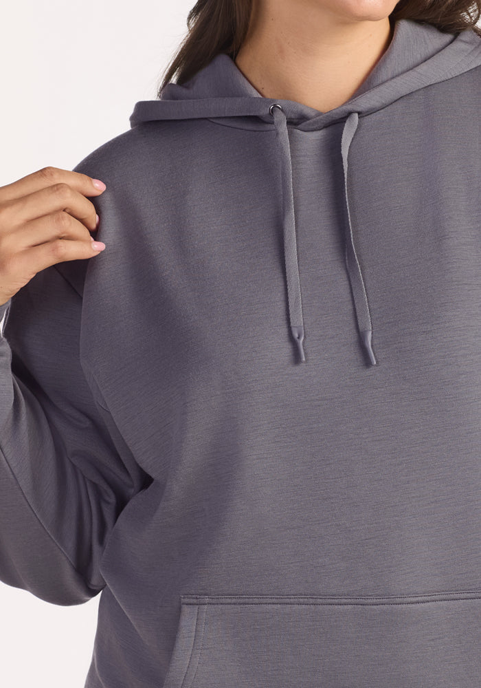 A woman, shown from shoulders to waist, wears the Woolx Avery Hoodie in Arctic Dusk—a loose-fitting gray Merino wool hoodie with drawstrings and a front pocket—holding the edge of her hood with one hand.
