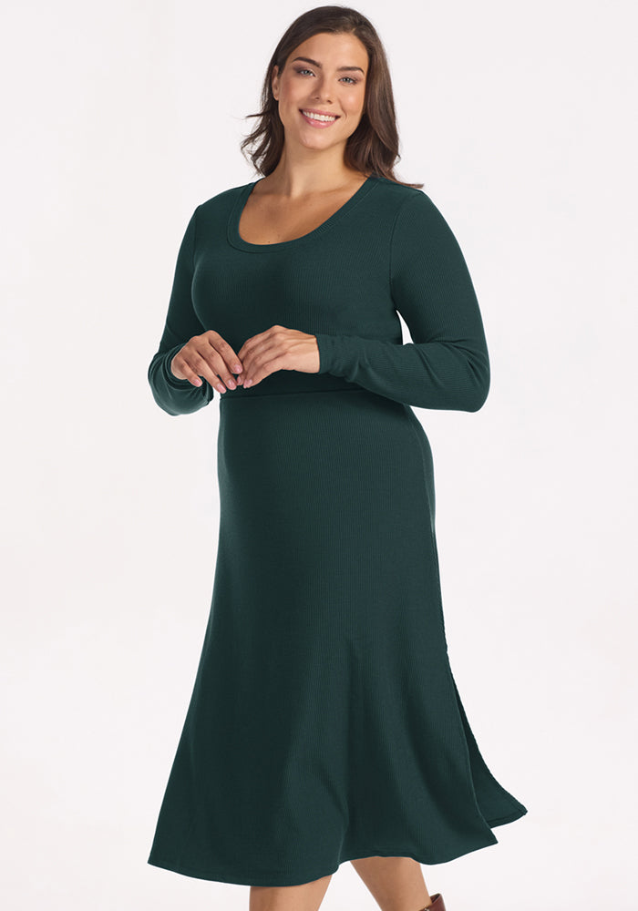 A woman with long brown hair smiles in the Woolx Caroline Ribbed Twirl Dress in Deep Timber, a long-sleeve ribbed merino style, while posing against a plain white background.