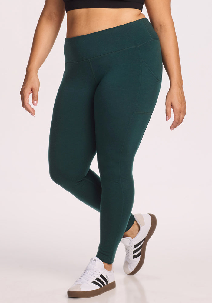 Wearing Woolx Piper Pocket Leggings in Deep Timber, a black crop top, and white Adidas sneakers, a person stands against a plain light background with only their body from shoulders down visible.