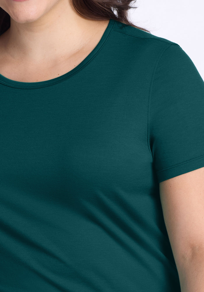 A close-up shows a person in the Woolx Liza Crew Neck Tee in Riverbank Green, featuring part of their face, neck, and upper arm against a plain background.