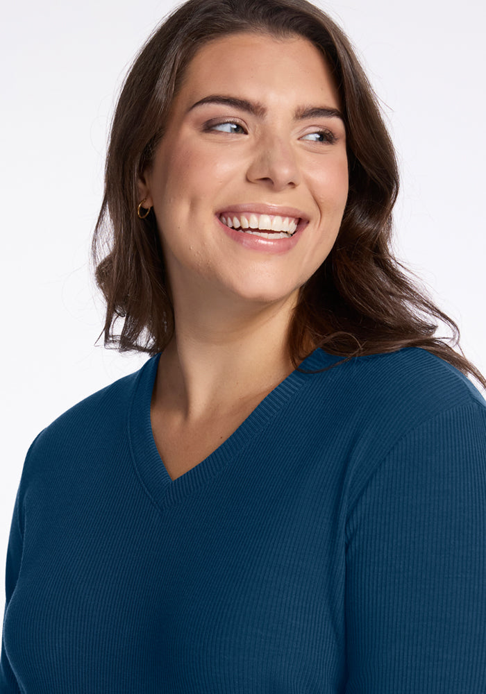 A woman with long brown hair smiles and looks off to the side against a white background, enjoying the temperature regulating comfort of the Woolx Evie Ribbed Tunic in Majolica Blue.