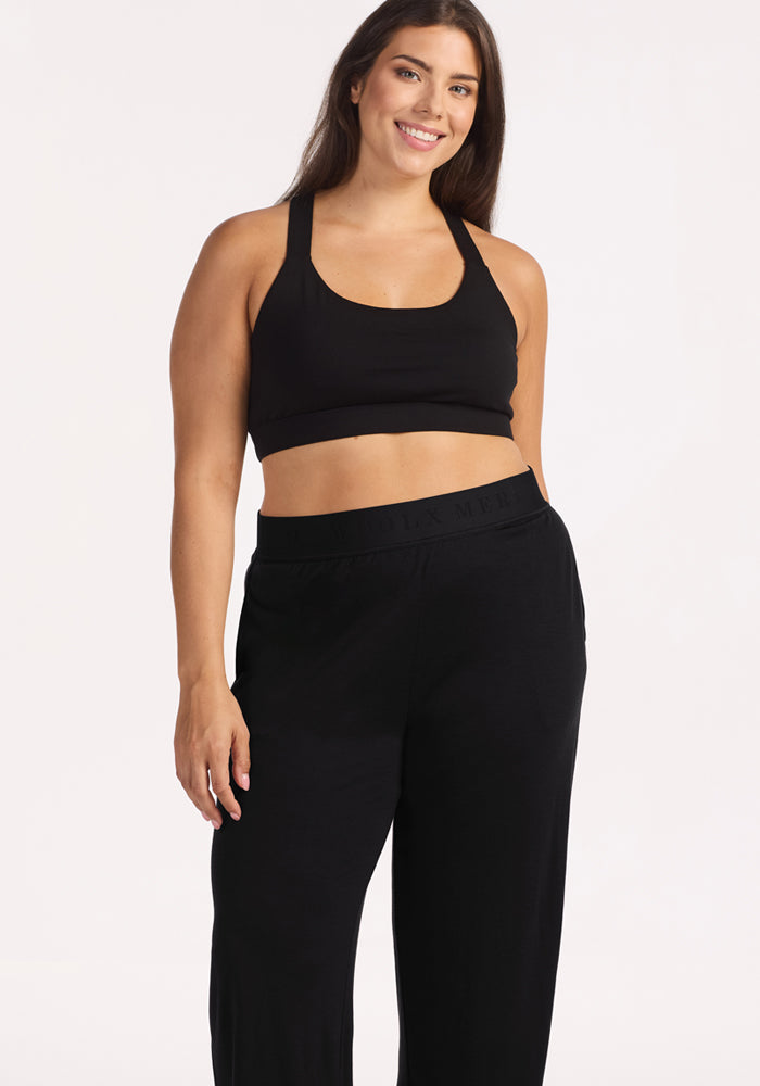 A woman with long brown hair smiles, wearing a black sports bra and Woolx Maya Lounge Pants in Black, standing against a plain light background.