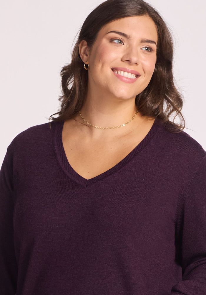 A woman with wavy brown hair smiles and looks to the side, wearing the Woolx Francesca Sweater in Deep Plum—a travel-friendly V-neck—paired with small hoop earrings and a delicate gold necklace against a light background.