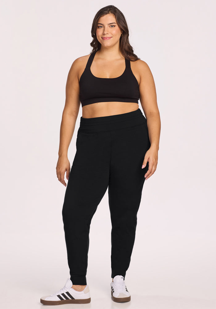 A woman wearing a black sports bra, Woolx Willa Joggers in Black, and white sneakers stands smiling against a plain light background.