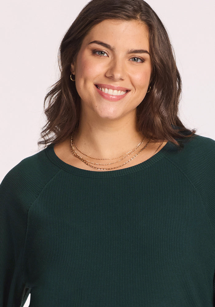 A woman with shoulder-length brown hair, wearing the Woolx Ainsley Sweater in Deep Timber and layered gold necklaces, smiles at the camera against a plain light background.
