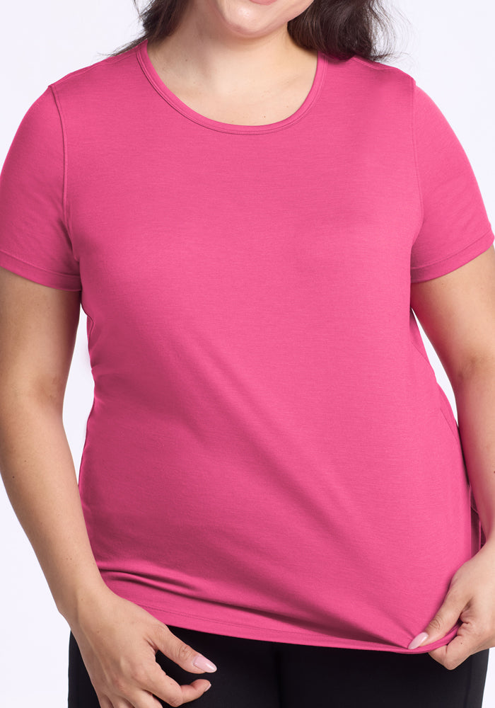 A woman stands against a light background, gently holding the hem of her Woolx Liza Crew Neck Tee in Raspberry Fizz. She pairs the lightweight performance top with black pants, and her face is partially out of frame.