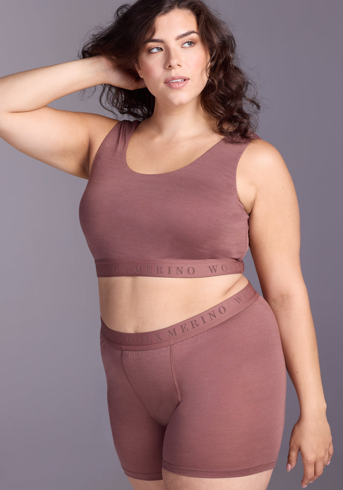 A woman with wavy hair poses confidently in Woolx's Elliott Boyshort - Nutmeg, a high-waisted antimicrobial underwear set, featuring a crop top and boyshorts, against a neutral gray background with her hand raised to her head.