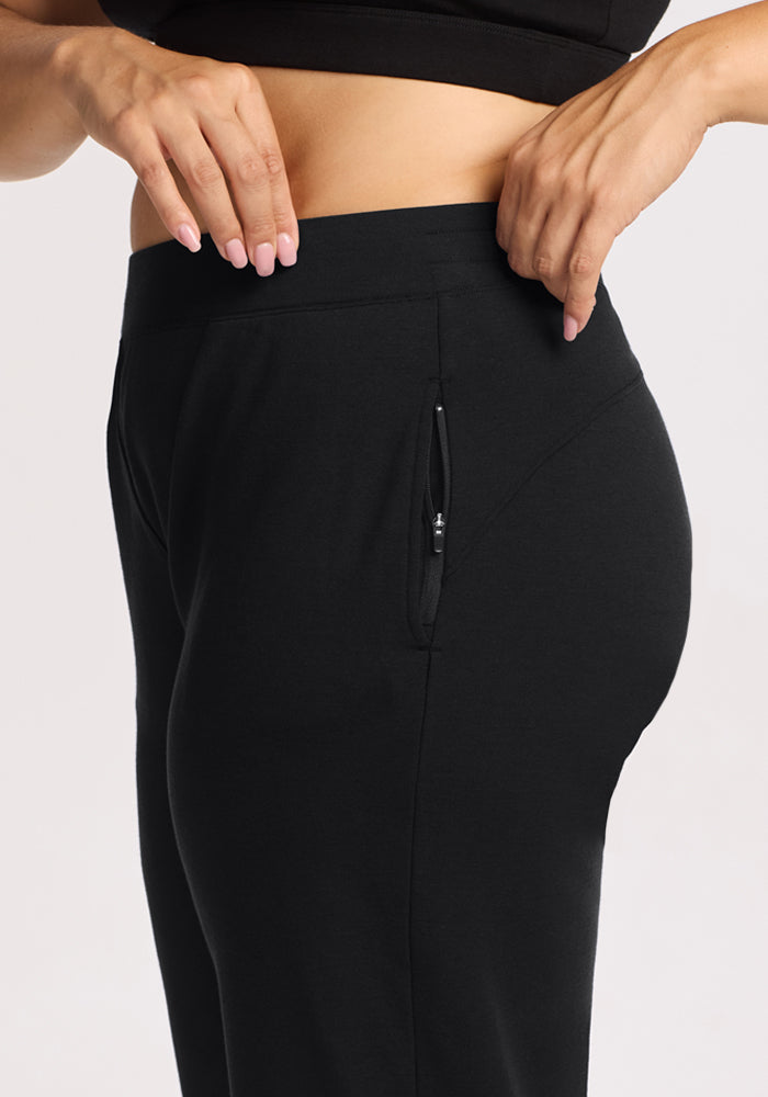 A close-up side view shows a person adjusting the waistband of Woolx Brynn Pants - Black, featuring a zippered pocket. The individual, with light skin and manicured nails, pairs them with a cropped black top.