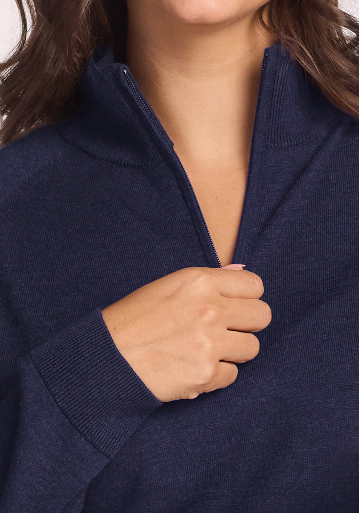 A person with wavy brown hair is shown from the shoulders up, zipping up the Woolx Collins Half Zip Sweater in Storm Cloud; only their lower face is visible.