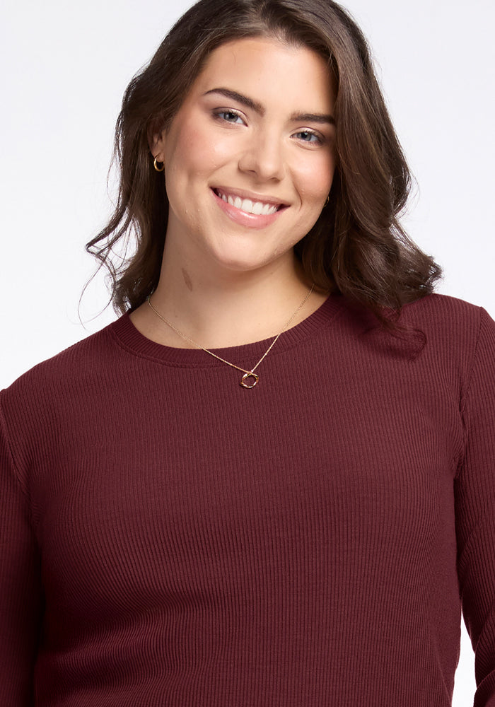 A woman with wavy brown hair smiles at the camera, wearing the Woolx Hadley Ribbed Crew in Toasted Garnet—perfect for lightweight layering. She pairs it with gold jewelry against a plain white background.