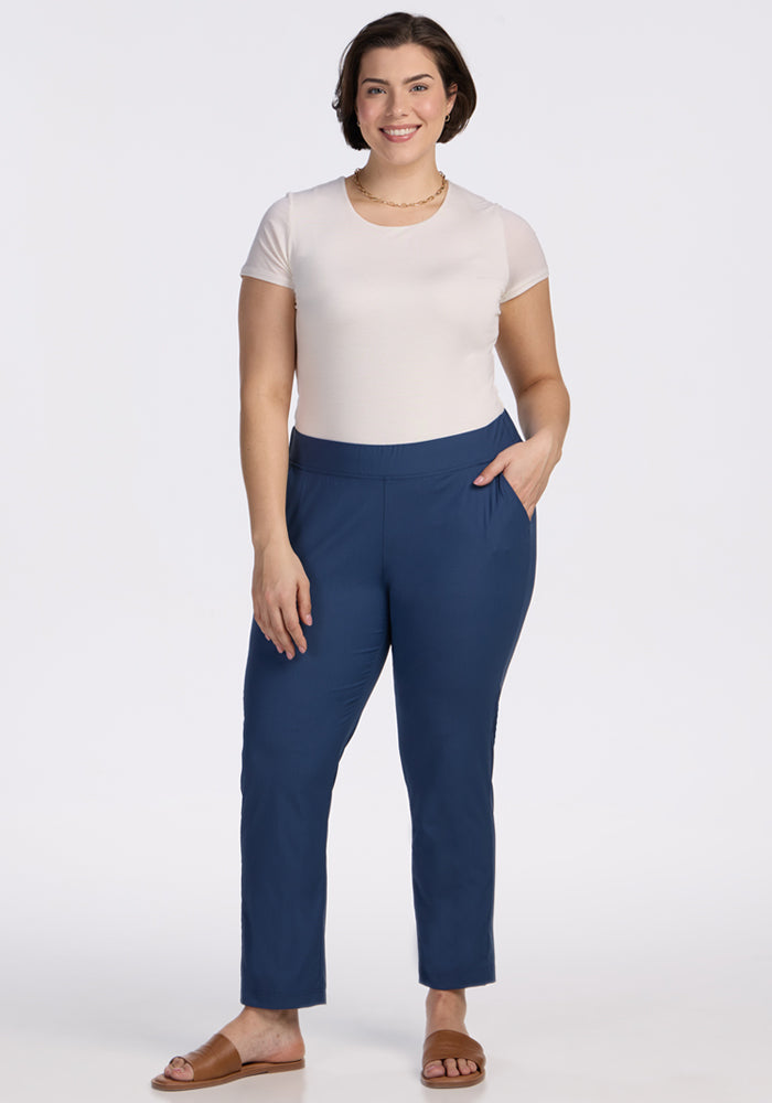 A woman stands facing forward, smiling, wearing a short-sleeve white t-shirt and Woolx Nicole Ankle Pants in Vintage Indigo with pockets, paired with brown sandals against a plain white background.