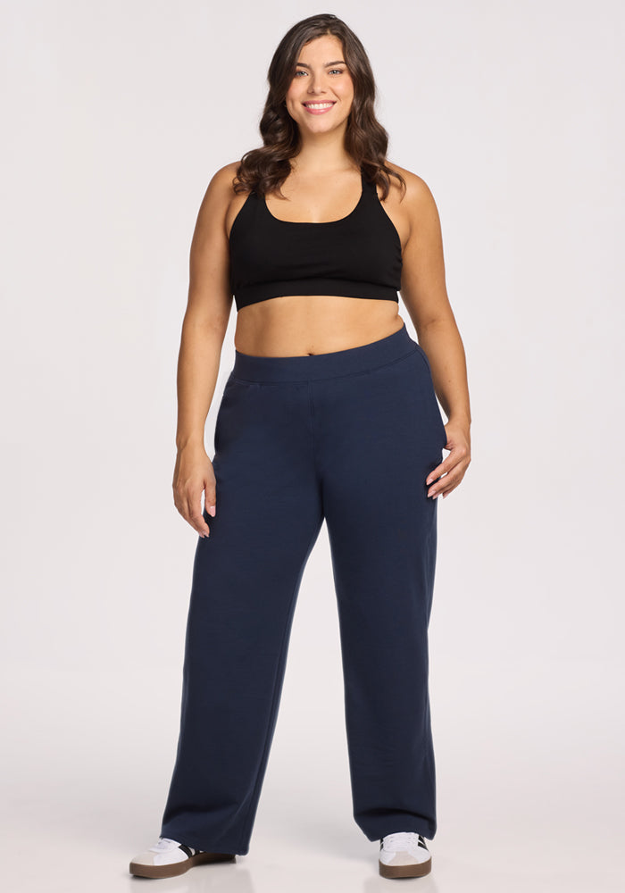A woman with long brown hair smiles in a black sports bra, Woolx Brynn Pants in Deep Navy, and white sneakers, set against a plain light background.