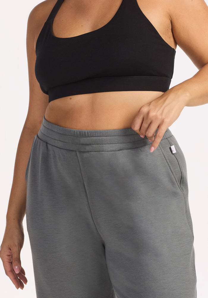 A woman models Woolx Parker Sweatpants in Sedona Sage, standing against a white background with one hand on the waistband, paired with a black sports bra.