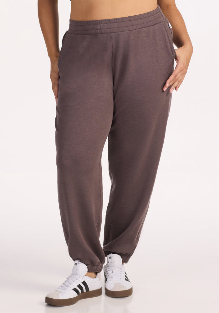 A person wearing Woolx Parker Sweatpants in Plum Truffle stands with one hand in their pocket, paired with white sneakers with black stripes, against a plain light background. 