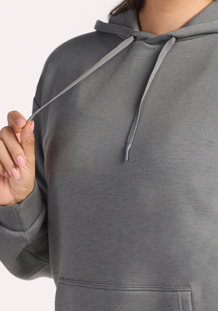 A person wearing the Woolx Avery Hoodie in Sedona Sage holds a drawstring near their neck, with only their upper body and hand visible against a neutral background.