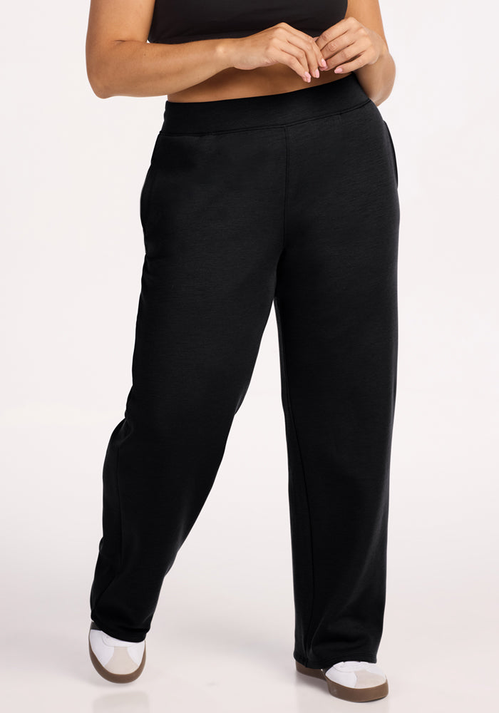 Only the lower torso and legs are visible as a person stands with hands clasped, wearing Woolx Bree Lounge Pants in black and white sneakers with tan soles.