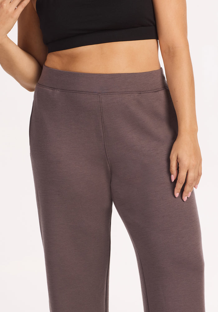 A person stands with one hand raised near their face and the other by their side, wearing a black crop top and Woolx Bree Lounge Pants in Plum Truffle. The image is cropped at the shoulders and thighs.