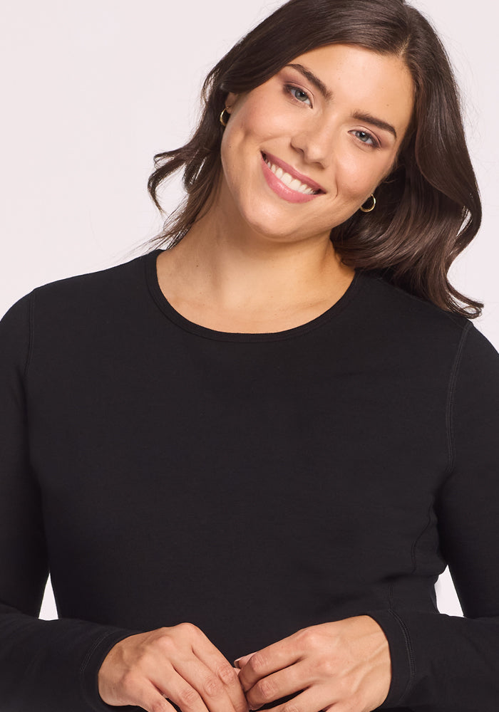 A woman with wavy dark hair wears the Woolx Elsa Long Sleeve in black and smiles warmly at the camera against a plain light background.