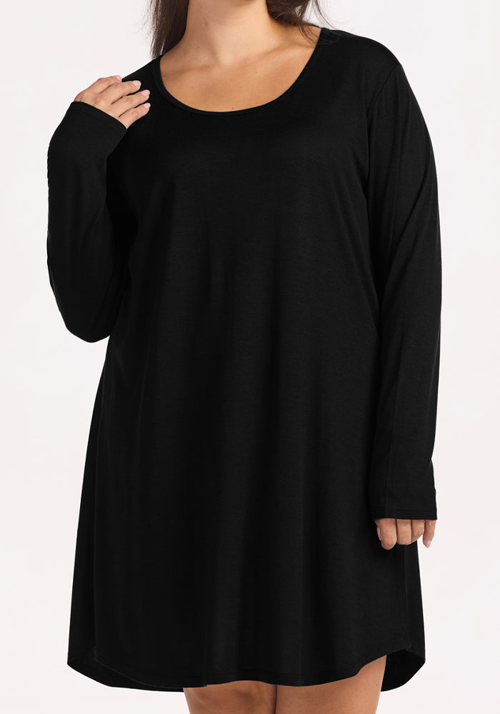 A woman wears the Woolx Lucille Long Sleeve Nightgown in black, featuring a loose fit, round neckline, and above-the-knee length. The background is plain and light-colored; her face is partially out of frame.