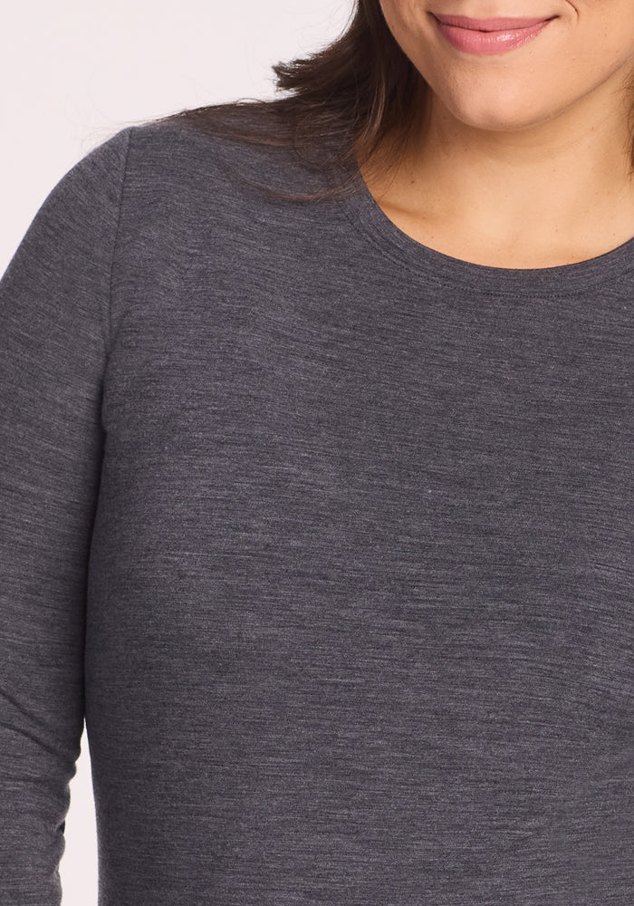 A woman smiles softly, wearing the Woolx Sadie Baselayer Top in Cinder Melange. The image features her upper torso and part of her face against a neutral background.
