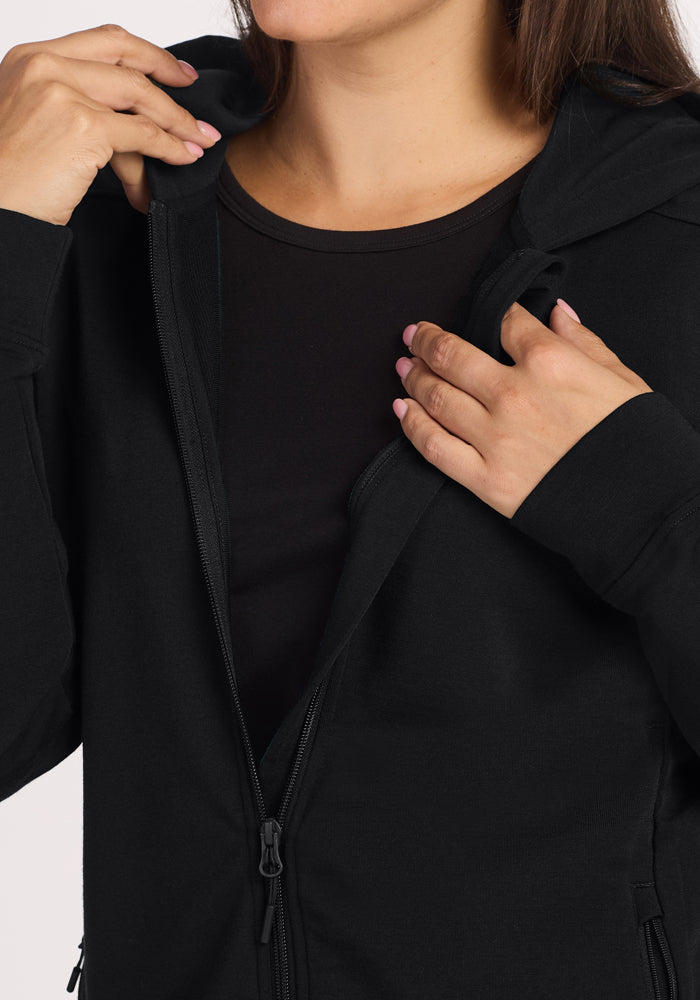 A woman is unzipping her Woolx Darby Zip Up Hoodie - Black, revealing a black shirt. Only the lower half of her face and hands with light pink painted nails are visible.