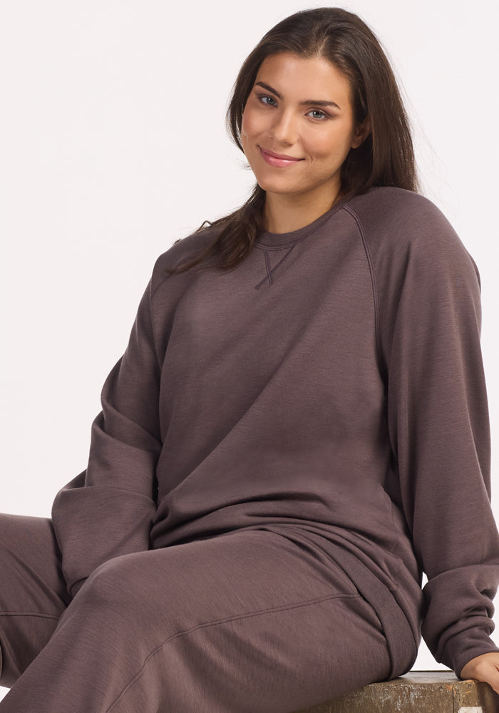 A woman with long brown hair wears the Woolx Bailey Crewneck Sweatshirt in Plum Truffle, featuring a recycled polyester exterior and Merino wool interior, as she sits on a wooden surface and smiles at the camera against a white background.