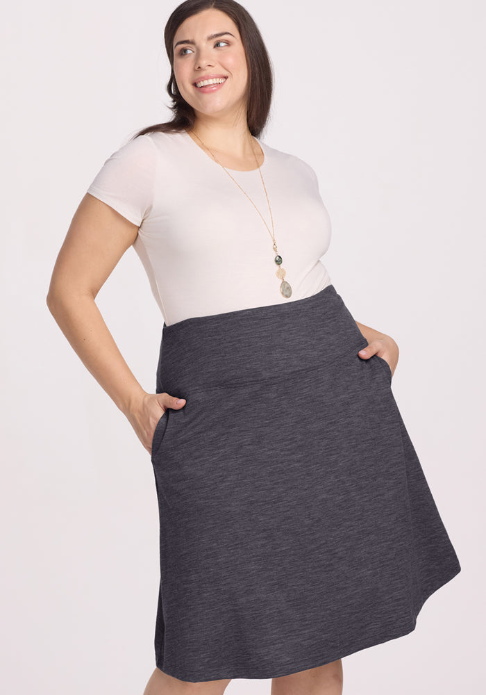 A smiling woman with long brown hair wears the Woolx Marilyn Flowy Midi Skirt in Cinder Melange, paired with a white short-sleeve top and a long pendant necklace, standing against a plain light background.