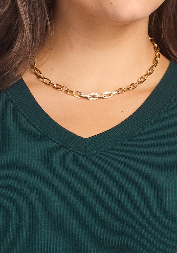 A woman wears the Woolx Evie Ribbed Tunic in Deep Timber, featuring a ribbed V-neck and paired with a chunky gold chain necklace. Only the lower half of her face, neck, and upper chest are visible.