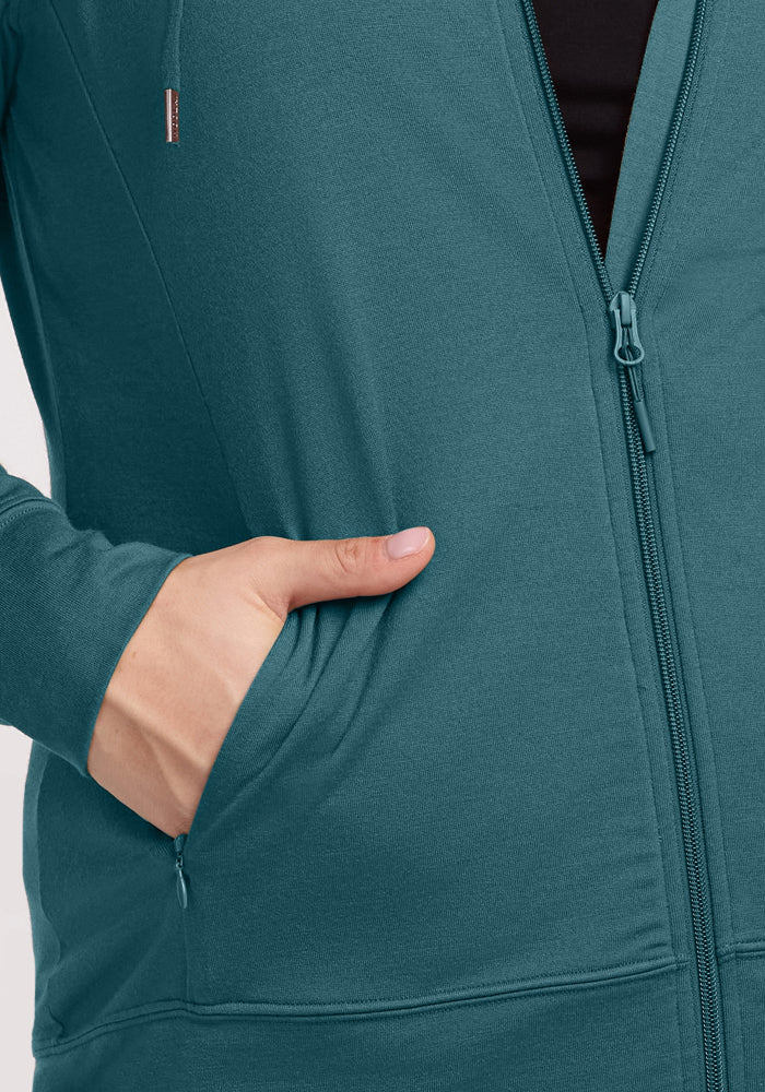 A person wears the Woolx Ryann Hoodie in Tidepool, left hand in the pocket and jacket partially unzipped to show a black shirt—an ideal travel essential.